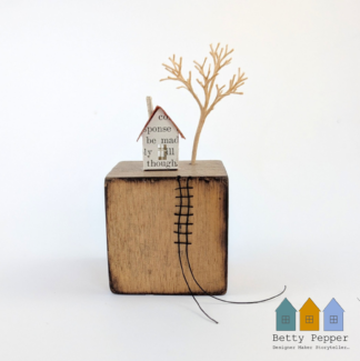Miniature paper houses handcrafted from faded vintage book covers and book pages, perched on a natural vintage child's wooden building block base with gently charred edges. A tiny thread ladder hangs down the side of the reel. By British artist Betty Pepper.