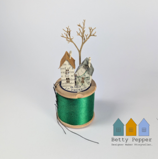 Miniature paper house handcrafted from repurposed book pages perched on a vintage wooden cotton reel wound with vibrant emerald green sewing thread creating a striking contrast between the old paper art and the rich green thread. A tiny thread ladder hangs down the side of the reel. By British artist Betty Pepper.