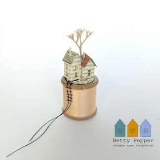 Miniature paper house handcrafted from repurposed book pages perched on a vintage wooden cotton reel wound with beautiful pale peachy pink thread. A tiny thread ladder hangs down the side of the reel. By British artist Betty Pepper.