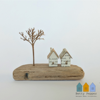 Miniature paper houses handcrafted from faded vintage book covers and book pages, perched on a natural driftwood base. This delicate sculpture features subtle, neutral tones and natural materials by British artist Betty Pepper.
