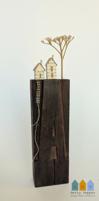 Miniature paper houses handcrafted from faded vintage book covers and book pages, perched on a vintage printer’s wooden letter ‘A’ block base. A tiny thread ladder in a contrast colour hangs down the side of the block. By British artist Betty Pepper.