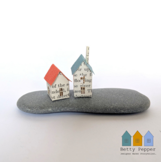 Miniature paper houses handcrafted from faded vintage book covers and book pages, perched on a natural pebble base. This delicate sculpture features subtle, neutral tones and natural materials by British artist Betty Pepper.