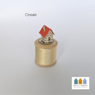 A vintage wooden cotton reel with cream thread. A tiny paper house sits on the reel it is made from an old book page and has an orange coloured roof. By Artist Betty.