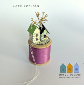 Miniature paper house handcrafted from repurposed book pages perched on a vintage wooden cotton reel wound with purple thread. A tiny thread ladder hangs down the side of the reel. By British artist Betty Pepper.
