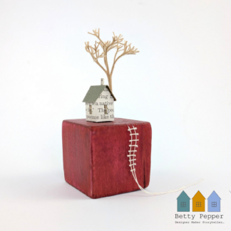 Miniature paper house handcrafted from faded vintage book covers and book pages, perched on a red vintage child's wooden building block base. A tiny thread ladder hangs down the side of the block. By British artist Betty Pepper.