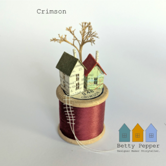 Miniature paper house handcrafted from repurposed book pages perched on a vintage wooden cotton reel wound with deep crimson thread. A tiny thread ladder hangs down the side of the reel. By British artist Betty Pepper.