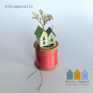 Miniature paper house handcrafted from repurposed book pages perched on a vintage wooden cotton reel wound with bright pink thread. A tiny thread ladder hangs down the side of the reel. By British artist Betty Pepper.