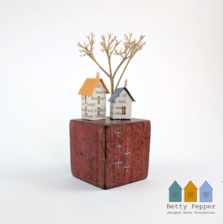 Miniature paper house handcrafted from faded vintage book covers and book pages, perched on a cherry red vintage child's wooden building block base. The block is embellished with duck egg blue cross stitches. By British artist Betty Pepper.
