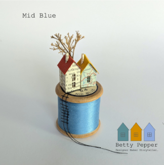 Miniature paper house handcrafted from repurposed book pages perched on a vintage wooden cotton reel wound with bright blue thread. A tiny thread ladder hangs down the side of the reel. By British artist Betty Pepper.