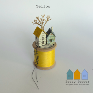 Miniature paper house handcrafted from repurposed book pages perched on a vintage wooden cotton reel wound with bright yellow thread. A tiny thread ladder hangs down the side of the reel. By British artist Betty Pepper.
